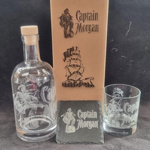 May include: A set of three Captain Morgan themed items: a glass bottle, a glass tumbler, and a black slate coaster. The bottle and tumbler are etched with a pirate ship and the Captain Morgan logo. The coaster features the Captain Morgan logo and the words "Captain Morgan".