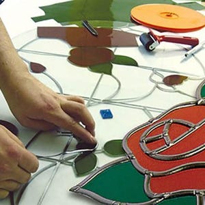 Op de afbeelding: Een glas-in-loodproject in uitvoering met rode en groene glasstukken verbonden door loodstrips. Het ontwerp bevat bloemmotieven met een rode roos en groene bladeren. Gereedschap omvat een zwarte marker, een kleine roller en een pen.