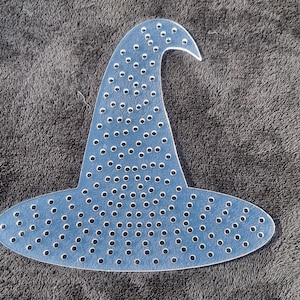May include: A light blue witch hat decoration with many small, evenly spaced holes. The hat has a curved brim and a pointed tip. The object is made of a flat, rigid material and is set against a dark grey, textured background.