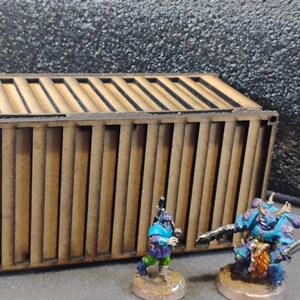 Może przedstawiać: Brązowy drewniany kontener wysyłkowy z linijką o długości 10 cm. Dwie miniaturowe figurki, jedna niebieska, a druga zielona, stoją przed kontenerem.