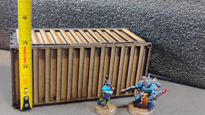 Może przedstawiać: Brązowy drewniany kontener wysyłkowy z linijką o długości 10 cm. Dwie miniaturowe figurki, jedna niebieska, a druga zielona, stoją przed kontenerem.