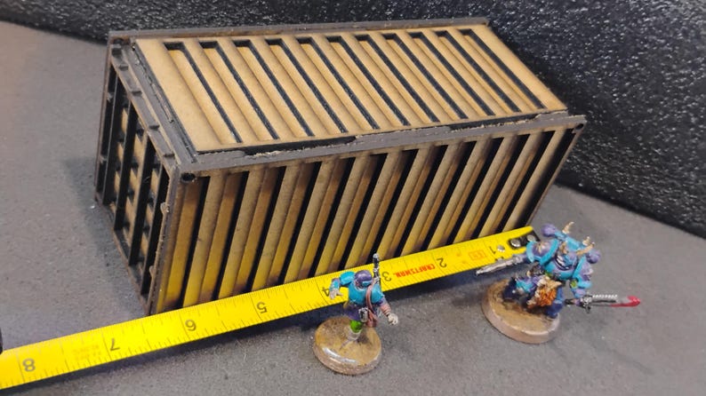 Może przedstawiać: Drewniany model kontenera transportowego z czarną obw&oacute;dką. Kontener ma około 30 cm długości i 10 cm szerokości. Kontener stoi na stole z ż&oacute;łtą miarką.