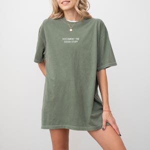 Könnte beinhalten: Olivgrünes übergroßes T-Shirt mit dem weißen Text "DOCUMENT THE GOOD STUFF". Das Shirt hat kurze Ärmel und eine lockere Passform. Das Model trägt eine weiße Halskette.