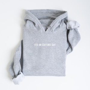 May include: A gray hoodie with the text "ITS AN EDITING DAY" in white. The hoodie is folded neatly on a white surface. The sleeves are cuffed, revealing a lighter gray interior.