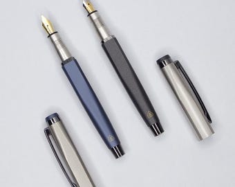 May include: Three fountain pens with different colored barrels: a dark gray pen, a light blue pen, and a silver pen. Each pen has a gold-colored nib and a small logo on the barrel.