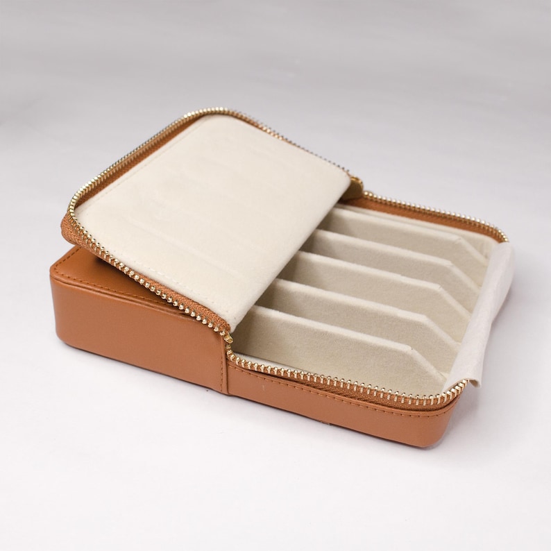 May include: A tan leather jewelry box with a zipper closure and multiple compartments for storing rings, earrings, or other small jewelry items.