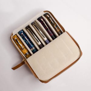 May include: A brown leather zippered case with a beige lining, open to reveal six colorful pens. The pens are arranged in a row, with the caps facing the viewer.