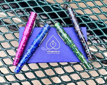 May include: Four fountain pens with different colorful marbled patterns, including pink, blue, green, and brown, are displayed on a blue box with the text "EPITOME PEN CO." The pens are arranged in a semi-circle on the box.