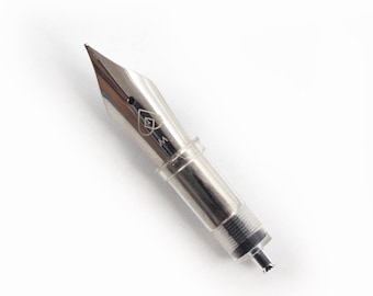 May include: A silver-toned fountain pen nib attached to a clear plastic feed and grip section. The metal nib features an engraved heart-shaped outline with a small diamond shape inside, positioned above a small circular breather hole.