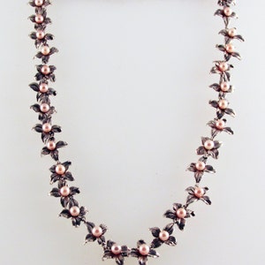 May include: A silver necklace with a delicate floral design and pink pearls. Each flower is accented with a single pearl, creating a feminine and elegant look.
