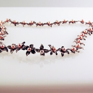 May include: A silver necklace with delicate flower-shaped charms and pink pearls. The necklace is laid out on a white surface.