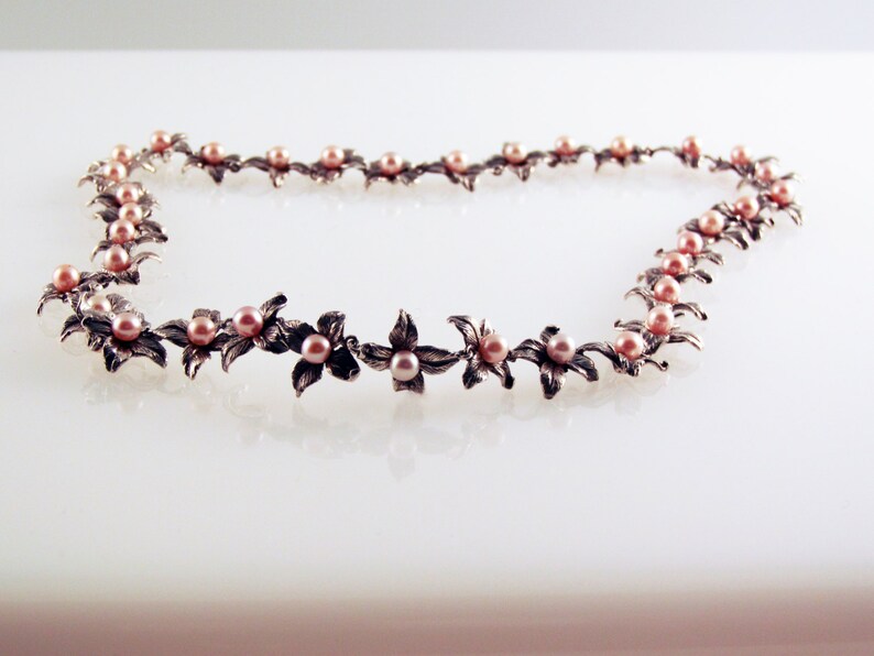 May include: A silver necklace with delicate flower-shaped charms and pink pearls. The necklace is laid out on a white surface.