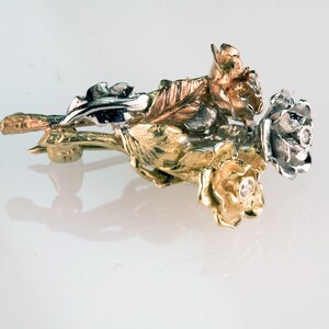 May include: A gold and silver brooch in the shape of three roses. The roses are detailed and have a realistic look. The brooch has a pin on the back for attaching to clothing.