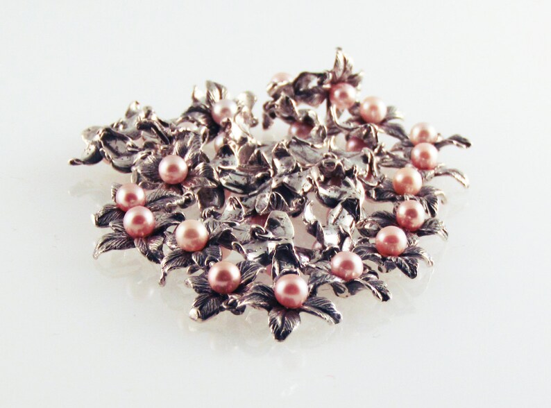 May include: A silver brooch with a floral design and pink pearls. The brooch is made of intricate metalwork and features a cluster of pearls in the center.