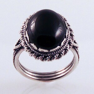 May include: A silver ring with a large, oval, black stone. The stone is set in a decorative, twisted rope-like frame. The ring has a triple-band design.