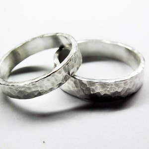 May include: Two silver hammered wedding bands, one slightly larger than the other, are stacked together on a white surface.
