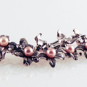 May include: A silver necklace with delicate floral designs and pink pearls. The necklace is made of metal and features a repeating pattern of flowers and pearls.