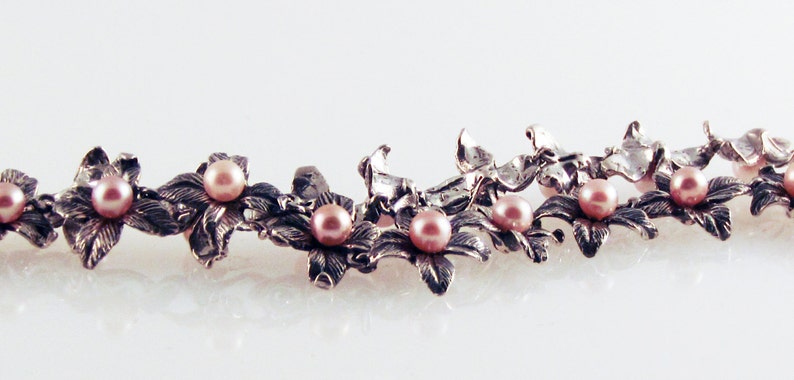 May include: A silver necklace with delicate floral designs and pink pearls. The necklace is made of metal and features a repeating pattern of flowers and pearls.