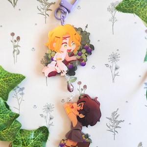 Op de afbeelding: Een sleutelhanger met twee cartoonfiguren, een met blond haar en de ander met bruin haar, beide houden paarse vruchten vast. De sleutelhanger heeft een paarse metalen sluiting.