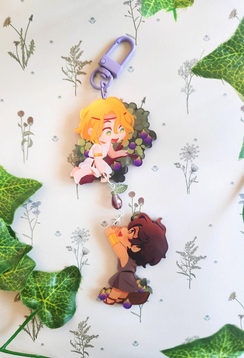 Op de afbeelding: Een sleutelhanger met twee cartoonfiguren, een met blond haar en de ander met bruin haar, beide houden paarse vruchten vast. De sleutelhanger heeft een paarse metalen sluiting.