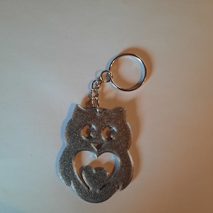 May include: A silver owl keychain with a heart cut out in the center. The owl is holding the heart in its wings.