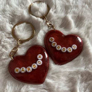 May include: Two heart-shaped keychains with the word "MAROON" spelled out in gold letters. The hearts are a deep red color with a glittery finish, attached to gold key rings.