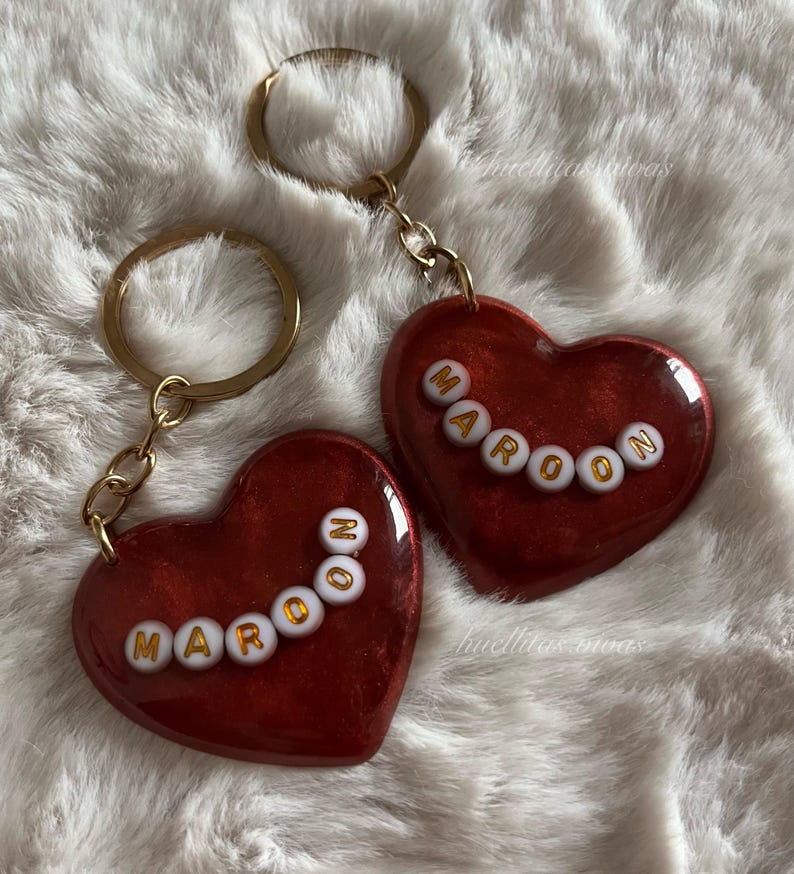 May include: Two heart-shaped keychains with the word "MAROON" spelled out in gold letters. The hearts are a deep red color with a glittery finish, attached to gold key rings.