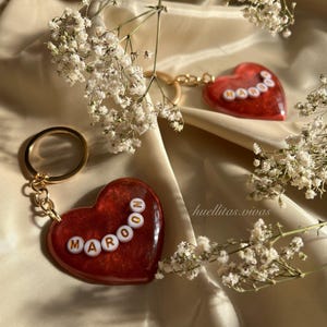 May include: Two heart-shaped keychains with gold-colored hardware. The keychains are maroon with white letter beads spelling "MAROON" and "OOOOO". The background is a beige fabric with white flowers.