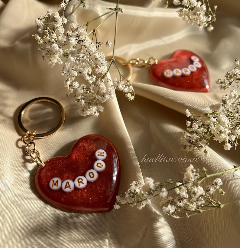 May include: Two heart-shaped keychains with gold-colored hardware. The keychains are maroon with white letter beads spelling "MAROON" and "OOOOO". The background is a beige fabric with white flowers.