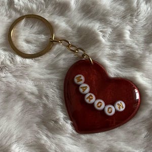 May include: Maroon heart-shaped keychain with gold-colored hardware. The word "MAROON" is spelled out in white and gold beads across the heart. The keychain is on a fluffy white surface.