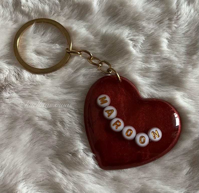 May include: Maroon heart-shaped keychain with gold-colored hardware. The word "MAROON" is spelled out in white and gold beads across the heart. The keychain is on a fluffy white surface.