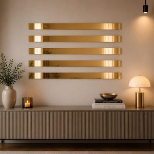 May include: A long, rectangular, light brown sideboard with a vertical ribbed texture, topped with a white lamp, books, and a plant. Above the sideboard, five horizontal, reflective gold-toned mirror strips are mounted on the wall in a parallel arrangement.