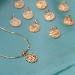 May include: A gold chain necklace with a small gold pendant featuring a zodiac symbol. The necklace is displayed on a turquoise blue fabric background. There are several other gold pendants with zodiac symbols scattered around the necklace.