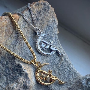 Puede incluir: Dos collares con colgantes de hada y luna creciente. Un collar es dorado y el otro es plateado. Ambos collares están sobre un fondo de roca.