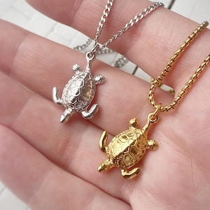 Peut inclure: Deux colliers avec pendentifs en forme de tortue. L'un est argenté, l'autre doré. Les pendentifs tortues ont des carapaces texturées et sont suspendus à de fines chaînes. Les colliers sont présentés sur un fond clair.