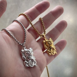 May include: Two necklaces with lucky cat pendants. One pendant is silver-colored, the other is gold-colored. Both pendants feature a cat with a raised paw and a coin. The necklaces have a chain link design.