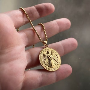 May include: A gold chain necklace with a round pendant featuring a guardian angel design. The pendant has the words "Guardian Angel Watch Over Me" inscribed around the edge.