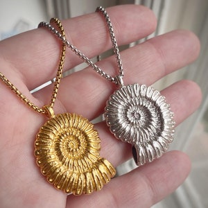 May include: Two gold and silver necklaces with a detailed ammonite pendant. The necklaces are on a hand, with the gold necklace on top and the silver necklace on the bottom.