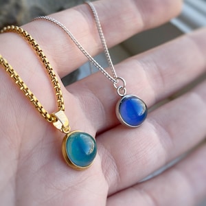 May include: Two pendant necklaces with round, blue gemstones. One has a gold chain and bezel, the other a silver chain and bezel. The gemstones have a gradient of colour.