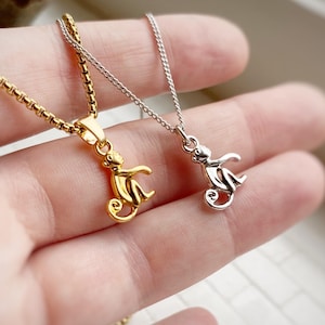 Puede incluir: Dos delicados collares con colgantes de mono. Uno es dorado y el otro plateado. Los colgantes de mono tienen un diseño estilizado. Los collares se muestran en una mano, mostrando los detalles y el tamaño de la joyería.
