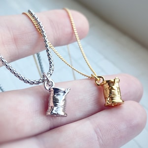May include: Two necklaces featuring spool pendants. One has a silver-coloured chain and pendant, the other a gold-coloured chain and pendant. The pendants are detailed with a textured design, resembling a thread spool.