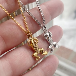 以下が含まれることがあります： カエルのペンダントが付いた2つのネックレス。1つは金色、もう1つは銀色です。どちらのネックレスも繊細なチェーンが付いています。カエルのペンダントは、カエルの特徴を示すテクスチャ加工された表面が特徴です。