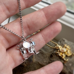 May include: A silver-colored necklace with a drum set pendant. The pendant features a detailed miniature drum kit with a round, iridescent center. The necklace is held in a hand, with a second gold-colored necklace visible in the background.