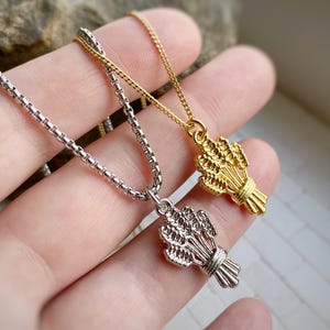 Könnte beinhalten: Zwei Halsketten mit Weizenähren-Anhängern. Eine Halskette hat eine silberfarbene Kette und einen Anhänger, die andere eine goldfarbene Kette und einen Anhänger. Die Anhänger sind mit einzelnen Weizenähren detailliert.