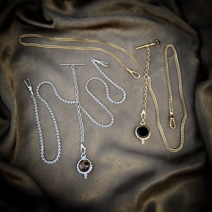 May include: Two pocket watch chains, one silver-toned and one gold-toned, each featuring a T-bar, a short drop chain with a circular pendant, and a longer chain section with a lobster clasp. The silver chain has a brown patterned stone pendant.