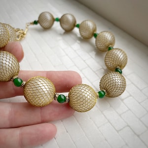 May include: A gold-toned necklace featuring large, round beads encased in a delicate mesh. The necklace is accented with small, vibrant green beads. The design is elegant and eye-catching, perfect for adding a touch of glamour to any outfit.