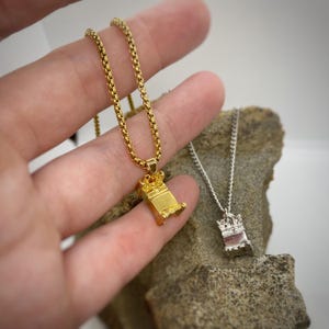 May include: Two gold necklaces with a small, detailed, box-shaped pendant. The pendant is topped with a crown. One necklace is on a hand, the other is on a rock.