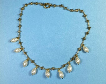Puede incluir: Un collar de oro con una cadena delicada y pequeñas cuentas nacaradas que cuelgan de él. El collar tiene un cierre de broche.