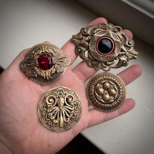 May include: Four ornate gold-toned brooches with intricate designs. One features a red gemstone, another has a black center with a red border, and two have a more abstract design.