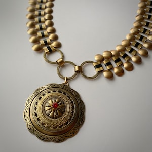 Peut inclure: Un collier vintage doré avec un grand pendentif orné. Le pendentif présente un bijou rouge central et des détails complexes. Le collier a une chaîne de disques dorés reliés avec des accents noirs, et est une pièce de déclaration.